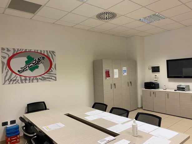 Sala riunioni con tavolo chiaro, sedie nere, armadi e monitor, pannello «Unità Operativa di Perugia» con giaguaro